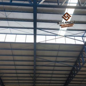 Detalhe do Lanternim Iluminação Solution instalado em galpão industrial, evidenciando a entrada de luz natural e a ventilação proporcionada pelo sistema. Detalhe do Lanternim Iluminação Solution em funcionamento, mostrando a entrada de luz natural e a ventilação no interior do galpão industrial.