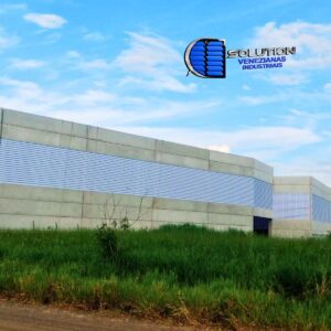 Fachada de galpão industrial em Santa Gertrudes, SP, com instalação de venezianas industriais Solution. A imagem mostra as aletas alinhadas garantindo ventilação permanente e iluminação natural em toda a extensão lateral da estrutura metálica.