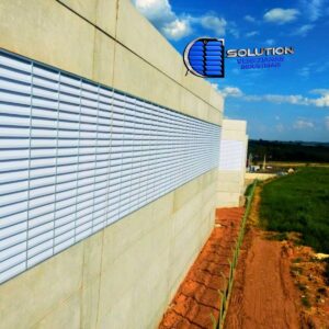 Fachada de galpão industrial com venezianas Solution instaladas em toda a extensão lateral, permitindo a entrada de luz solar difusa e a circulação constante de ar fresco no ambiente fabril.
