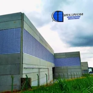 Fachada lateral de galpão industrial com sistema de venezianas industriais Solution instaladas em toda a extensão. A imagem demonstra a integração das venezianas com a estrutura metálica, permitindo a entrada de luz natural e a circulação constante de ar fresco no ambiente fabril.