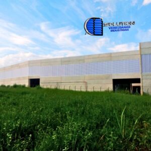 Fachada de galpão industrial com venezianas Solution instaladas em toda a extensão lateral. A imagem destaca as aletas translúcidas que permitem a entrada de iluminação natural e a circulação constante de ar no ambiente fabril.