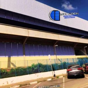 Fachada de galpão industrial com venezianas em policarbonato alveolar branco translúcido, mostrando a entrada difusa de luz natural e a estrutura metálica de suporte.