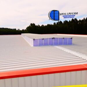 Vista detalhada de um lanternim industrial equipado com venezianas Solution em policarbonato branco translúcido. A imagem mostra a estrutura elevada no topo do telhado, permitindo a saída do ar quente e a entrada de luz solar difusa para o interior do galpão.