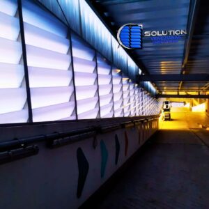 O policarbonato utilizado possui propriedades de difusão luminosa que otimizam a claridade interna, reduzindo a necessidade de iluminação artificial em grandes áreas cobertas. No aspecto térmico e de qualidade do ar, o design das aletas Solution permite uma taxa de renovação de ar (vazão) calculada para evitar o superaquecimento do pavimento e o acúmulo de monóxido de carbono proveniente dos veículos. A estrutura de fixação foi projetada para resistir a altas cargas de vento, comuns em áreas abertas de aeroportos, garantindo longevidade e baixa manutenção ao empreendimento.Destaques da Obra:Cliente: Estacionamento Pare BemLocal: Aeroporto (Paraná)Material: Policarbonato Branco Leitoso com Proteção UVFunção: Ventilação permanente e iluminação zenital/lateral para grandes vãos.