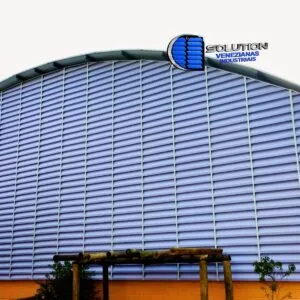 Vista de ginásio poliesportivo com venezianas industriais Solution fabricadas em arco. As aletas são de policarbonato alveolar branco leitoso translúcido, acompanhando a curvatura da cobertura para garantir ventilação lateral e iluminação zenital difusa