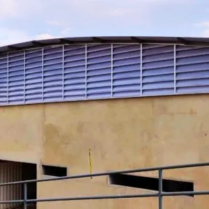 Fachada curva de ginásio poliesportivo com venezianas industriais Solution fabricadas em arco. As aletas são de policarbonato alveolar branco leitoso translúcido, permitindo a entrada de luz natural suave e ventilação permanente na arena esportiva.