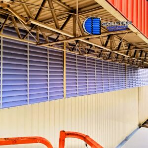 Fachada de indústria de alimentos com venezianas Solution em policarbonato branco leitoso translúcido. A imagem mostra as aletas instaladas em galpão industrial, proporcionando iluminação natural difusa e ventilação permanente para controle de temperatura interna.