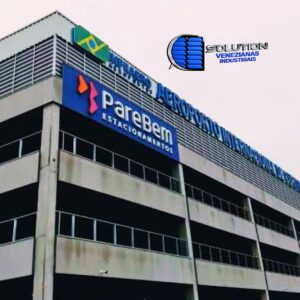 Fachada do estacionamento Pare Bem no Aeroporto do Paraná, apresentando venezianas industriais Solution em policarbonato branco leitoso translúcido. A imagem mostra a estrutura de fechamento lateral que garante ventilação constante e iluminação natural suave para a área de veículos.