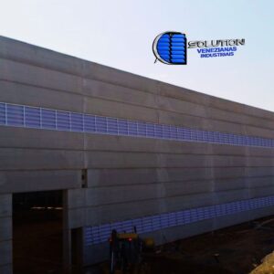 Fachada de galpão industrial com venezianas Solution em policarbonato branco leitoso translúcido. A imagem mostra a extensão lateral do galpão iluminada pela luz natural difusa, com aletas sobrepostas que garantem ventilação permanente e proteção contra chuva