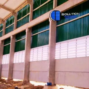 Fachada de galpão industrial com venezianas Solution em policarbonato branco leitoso translúcido. A imagem mostra as lâminas fixas instaladas em grandes vãos laterais, permitindo a passagem de luz solar filtrada e garantindo a ventilação natural do ambiente fabril.