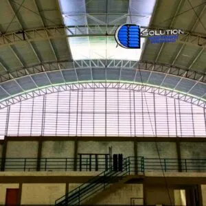 Fachada curva de ginásio poliesportivo com venezianas Solution em policarbonato alveolar branco leitoso. A imagem mostra a instalação em arco acompanhando a curvatura da cobertura, com aletas translúcidas que difundem a luz solar de forma homogênea.