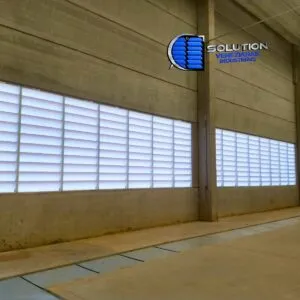Vista interna e externa de galpão industrial com fechamento lateral em venezianas Solution. A imagem demonstra a entrada abundante de luz natural, preenchendo o espaço de trabalho e reduzindo a dependência de iluminação elétrica durante o dia.