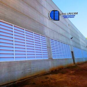 Fechamento lateral de galpão industrial com venezianas Solution. O sistema utiliza aletas translúcidas para maximizar a entrada de luz natural e aberturas estratégicas para ventilação permanente, eliminando a necessidade de iluminação artificial diurna.